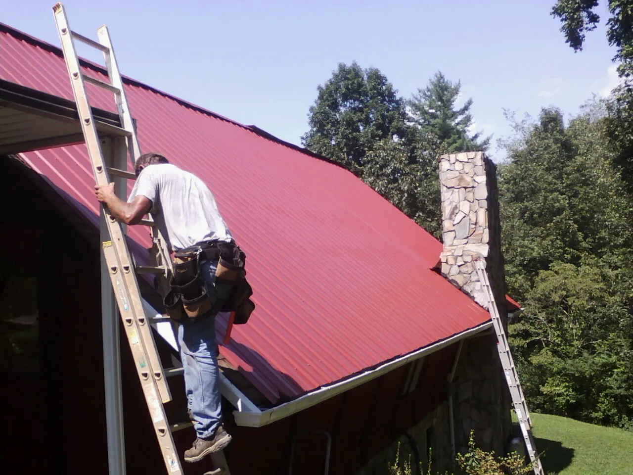 Roofer on a ladder beside a red metal roof and stone chimney — Soffit & Fascia Repair by Roofing Brothers in East Northport