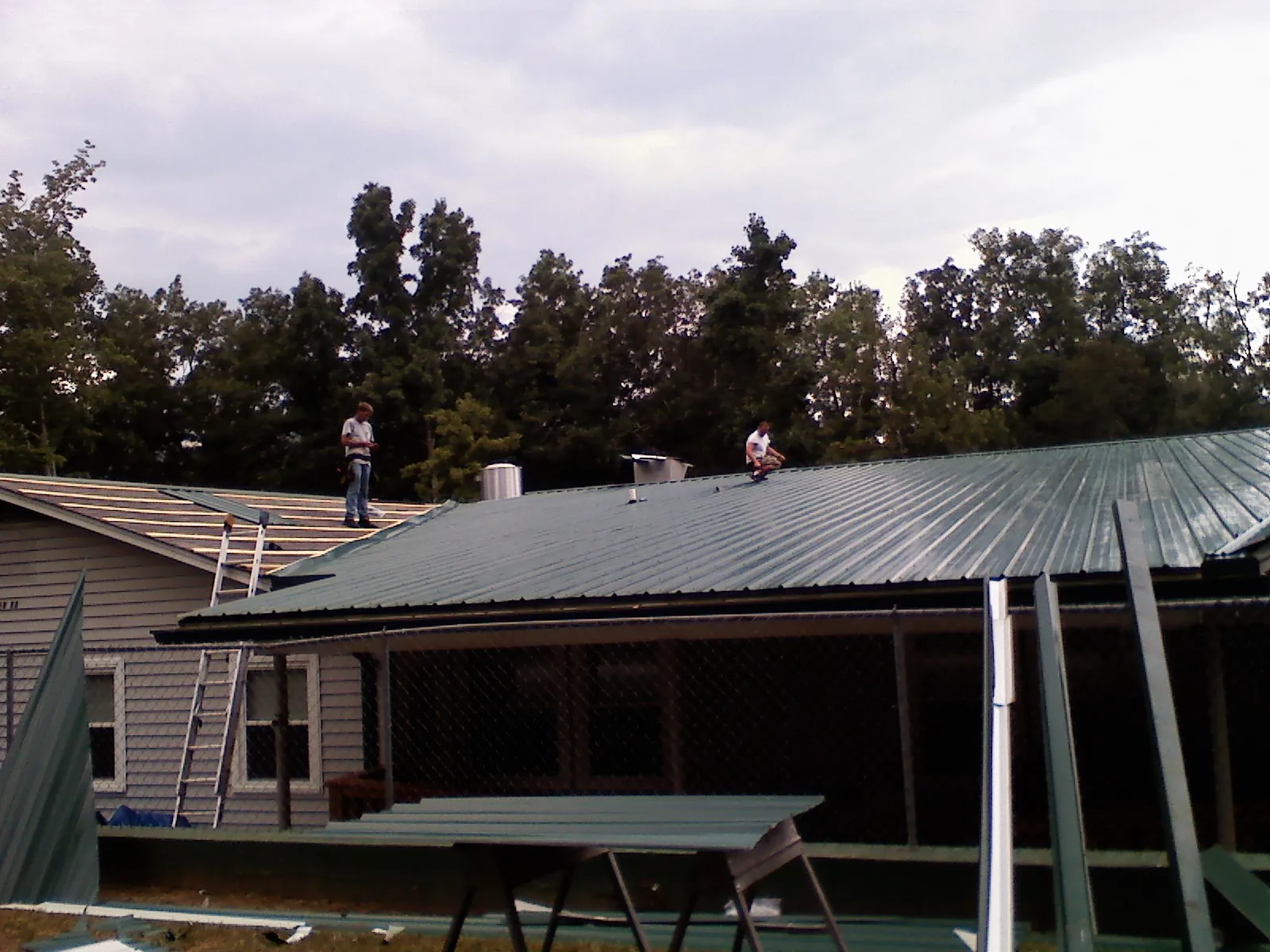 Two roofers installing green metal panels above a covered porch in East Northport